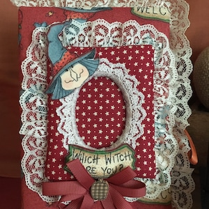 May include: A handmade Halloween-themed journal with a red star-patterned cover, adorned with a witch illustration, lace trim, and a ribbon bow. The text "WHICH WITCH ARE YOU?" is visible on a small sign. The journal is a unique, decorative item.