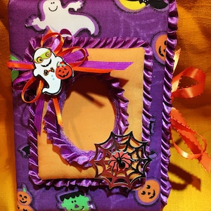 May include: A handmade Halloween-themed journal with a purple fabric cover featuring ghost and jack-o'-lantern patterns. It has an orange frame, a ghost embellishment, and a silver spiderweb decoration. The journal is tied with orange ribbon.
