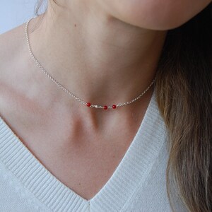 May include: A delicate silver necklace featuring three small, vibrant red beads. The necklace is displayed against a white sweater, highlighting the contrast between the silver chain and the red beads. A simple and elegant piece of jewelry.