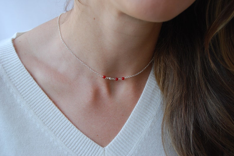 May include: A delicate silver necklace featuring three small, vibrant red beads. The necklace is displayed against a white sweater, highlighting the contrast between the silver chain and the red beads. A simple and elegant piece of jewelry.