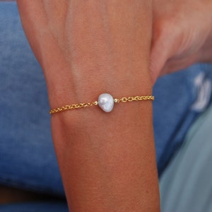 May include: A delicate gold chain bracelet with a single, off-white baroque pearl pendant.