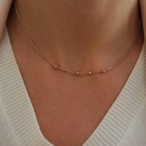 Puede incluir: Delicado collar dorado con pequeñas piedras preciosas rosas y cuentas doradas. El collar se lleva con un suéter blanco y una blazer rosa claro, creando un look suave y elegante. Una pieza de joyería sencilla y con estilo.