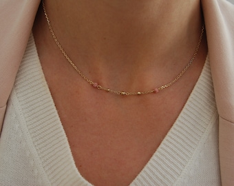 May include: Delicate gold necklace featuring small pink gemstones and gold beads. The necklace is worn against a white sweater and a light pink blazer, creating a soft and elegant look. A simple and stylish piece of jewelry.
