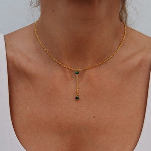 May include: A gold chain necklace with a delicate drop pendant featuring two small green gemstones.