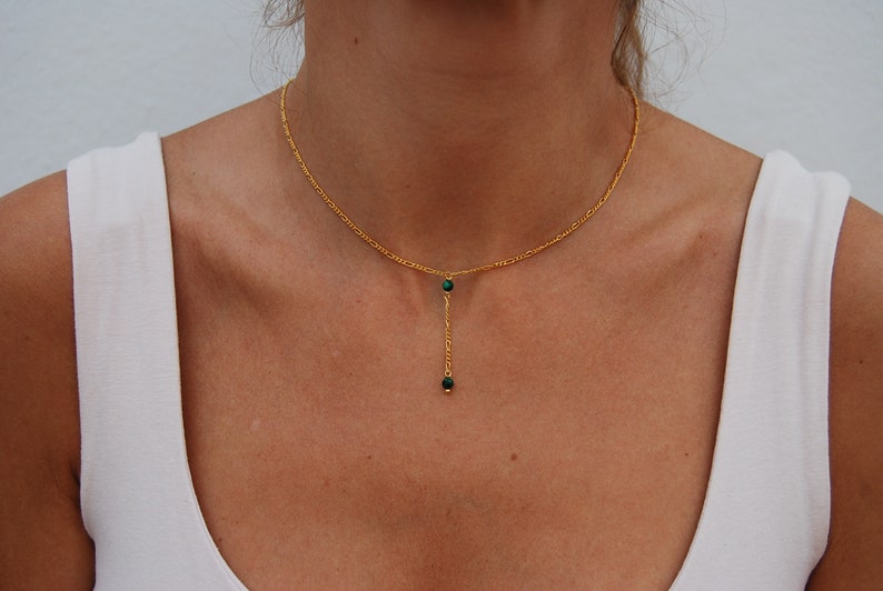 May include: A gold chain necklace with a delicate drop pendant featuring two small green gemstones.