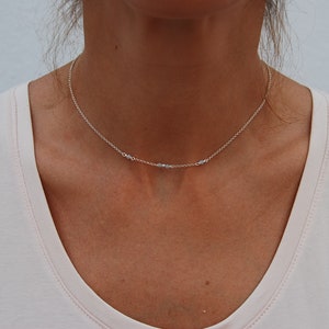 May include: A delicate silver chain necklace with small, clear beads. The necklace is worn by a person with a light pink shirt.