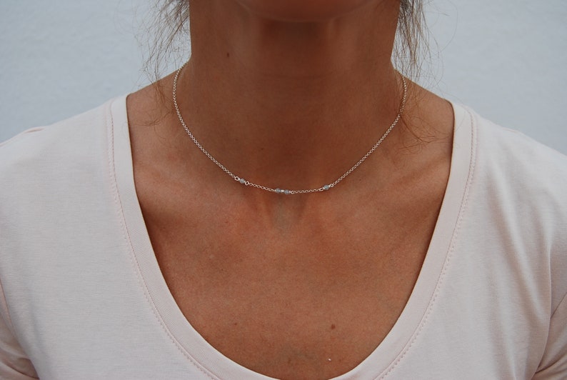 May include: A delicate silver chain necklace with small, clear beads. The necklace is worn by a person with a light pink shirt.