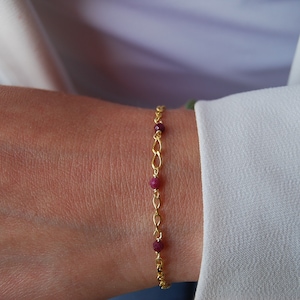 Può includere: Un braccialetto a catena dorata con piccole perle di pietre preziose rosse.