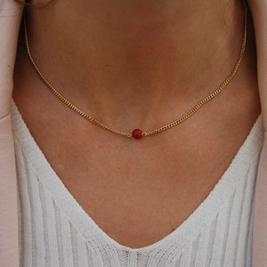 Puede incluir: Un collar de cadena dorada con un colgante de cuentas rojas.