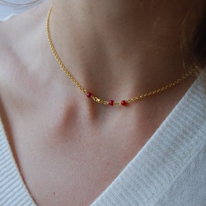 May include: A delicate gold-colored necklace featuring a fine chain and three small, vibrant red beads. The necklace is worn around the neck, creating a simple yet elegant look. The jewelry is a subtle accessory.