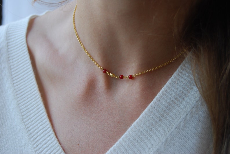 May include: A delicate gold-colored necklace featuring a fine chain and three small, vibrant red beads. The necklace is worn around the neck, creating a simple yet elegant look. The jewelry is a subtle accessory.
