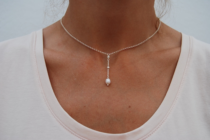 Puede incluir: Un collar de cadena de plata con un colgante de perla &uacute;nico. La cadena es delicada y la perla es peque&ntilde;a y blanca.