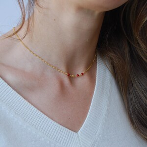 May include: A delicate gold necklace featuring a fine chain and a series of small, vibrant red beads. The necklace is shown against a white sweater, highlighting the jewelry's simple elegance and color contrast.