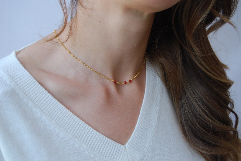 May include: A delicate gold necklace featuring a fine chain and a series of small, vibrant red beads. The necklace is shown against a white sweater, highlighting the jewelry's simple elegance and color contrast.