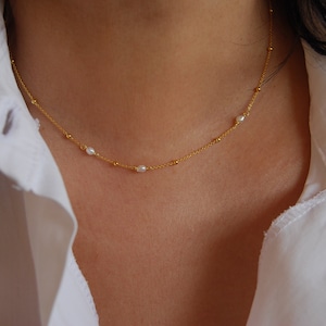 May include: A delicate gold chain necklace with small pearl accents. The necklace is worn by a person with a white shirt.