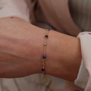 Puede incluir: Un brazalete de cadena dorada con pequeñas cuentas azules. El brazalete está en la muñeca de una persona.