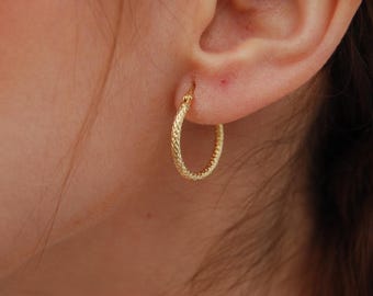 May include: A close-up of a gold hoop earring with a textured, woven design. The earring is worn on an ear, and the background is blurred. The earring is a classic jewelry piece.