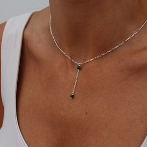 May include: A silver chain necklace with a delicate drop pendant featuring two small, dark green gemstones.