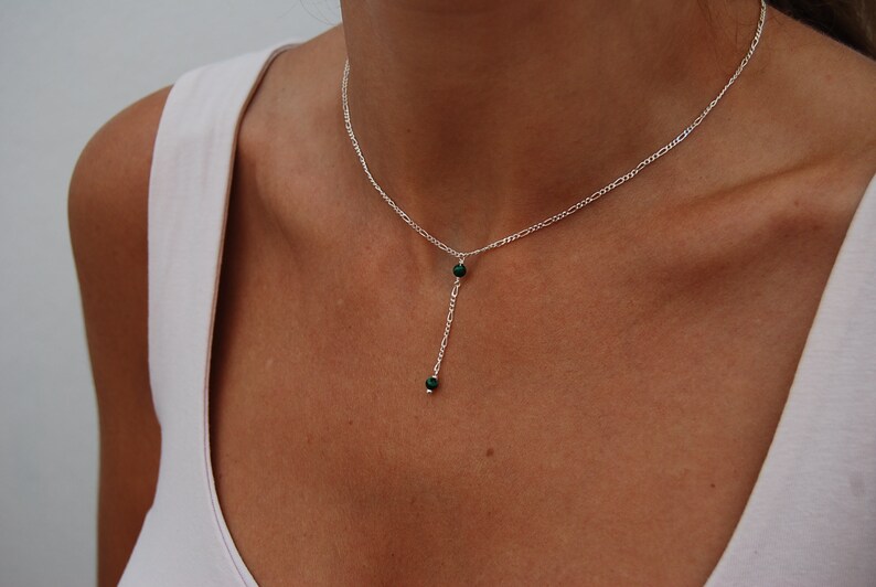 May include: A silver chain necklace with a delicate drop pendant featuring two small, dark green gemstones.