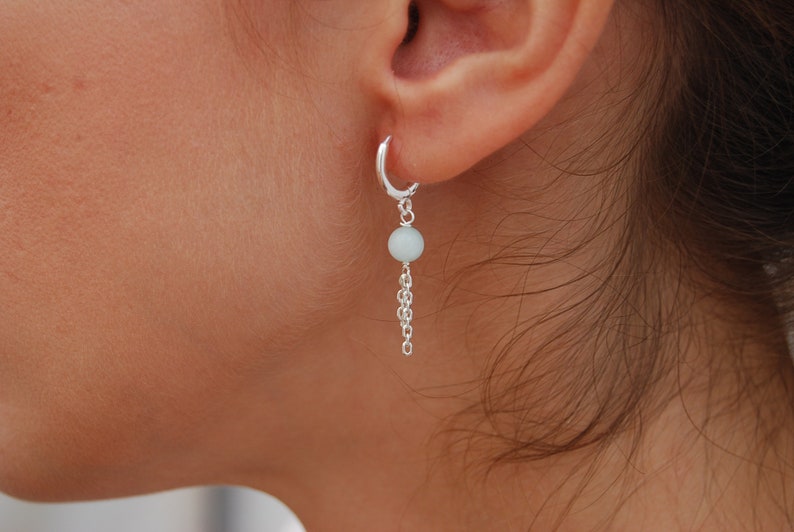 Pode incluir: Brincos de argola de prata com uma gema azul claro pendurada em uma corrente de prata.
