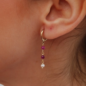 Könnte beinhalten: Goldene Creolen mit einer baumelnden Kette aus lila und weißen Perlen.