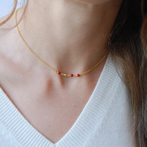 May include: A delicate gold necklace featuring a thin chain and a series of small, vibrant red beads. The necklace is displayed against a white background, highlighting its simple elegance. This piece is a stylish accessory.