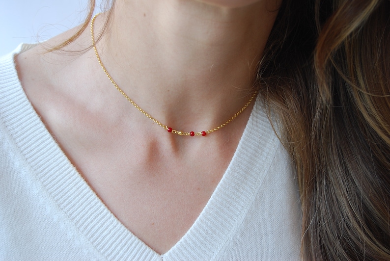 May include: A delicate gold necklace featuring a thin chain and a series of small, vibrant red beads. The necklace is displayed against a white background, highlighting its simple elegance. This piece is a stylish accessory.