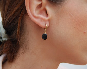 May include: Gold-colored hoop earrings with a dangling black oval stone. The earrings are shown on a person's ear, highlighting the contrast between the gold metal and the dark stone. A simple and elegant jewelry piece.