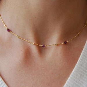 Op de afbeelding: Een delicate gouden ketting met kleine paarse edelstenen. De ketting wordt gedragen door een persoon.