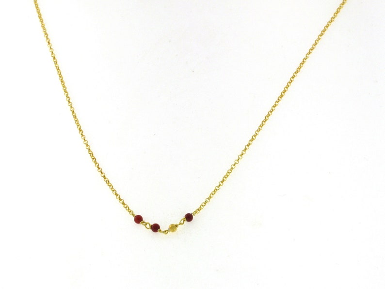May include: A delicate gold-colored necklace featuring a series of small, round, red beads and a single gold-colored bead. The beads are strung along a fine chain, creating a subtle and elegant design. The necklace is displayed against a white background.