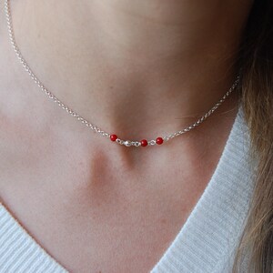 May include: A delicate silver necklace featuring a series of small, vibrant red beads interspersed with silver beads. The necklace is displayed on a person's neck, showcasing its simple yet elegant design. The chain is approximately 16 inches long.