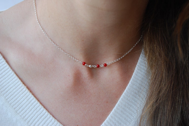 May include: A delicate silver necklace featuring a series of small, vibrant red beads interspersed with silver beads. The necklace is displayed on a person's neck, showcasing its simple yet elegant design. The chain is approximately 16 inches long.