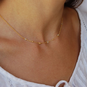 Puede incluir: Un delicado collar de cadena dorada con pequeñas cuentas transparentes. El collar lo lleva una persona con una camisa blanca.