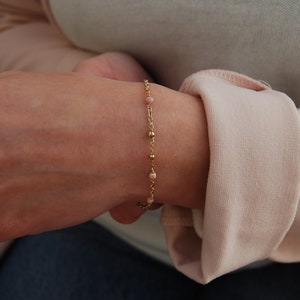 May include: A gold chain bracelet with small pink beads. The bracelet is on a person's wrist.