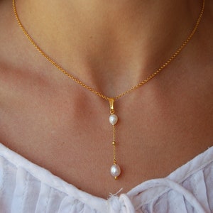 May include: A delicate gold chain necklace with a single pearl pendant and two smaller pearls dangling below. The necklace is worn by a person with a white shirt.
