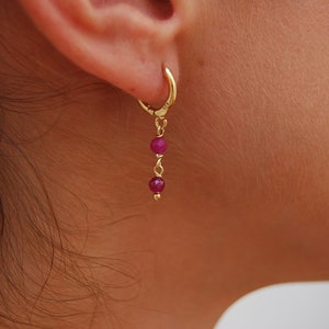 Peut inclure: Boucles d'oreilles créoles en or avec une chaîne pendante et trois petites pierres rondes roses.