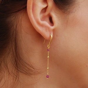 Peut inclure: Boucles d'oreilles pendantes dorées avec une petite pierre précieuse rose au bas de la chaîne.