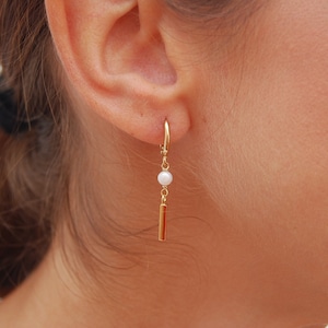 May include: Gold hoop earrings with a dangling pearl and a gold bar charm.
