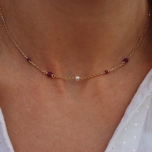 Op de afbeelding: Een gouden ketting met kleine rode edelstenen en een enkele witte parel hanger.