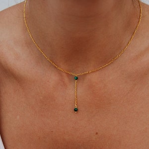 May include: A gold chain necklace with a delicate drop pendant featuring two small green gemstones.