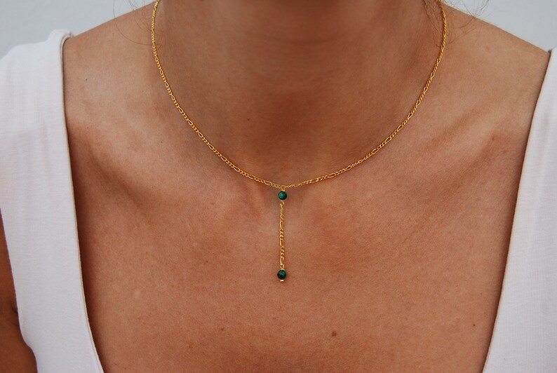 May include: A gold chain necklace with a delicate drop pendant featuring two small green gemstones.