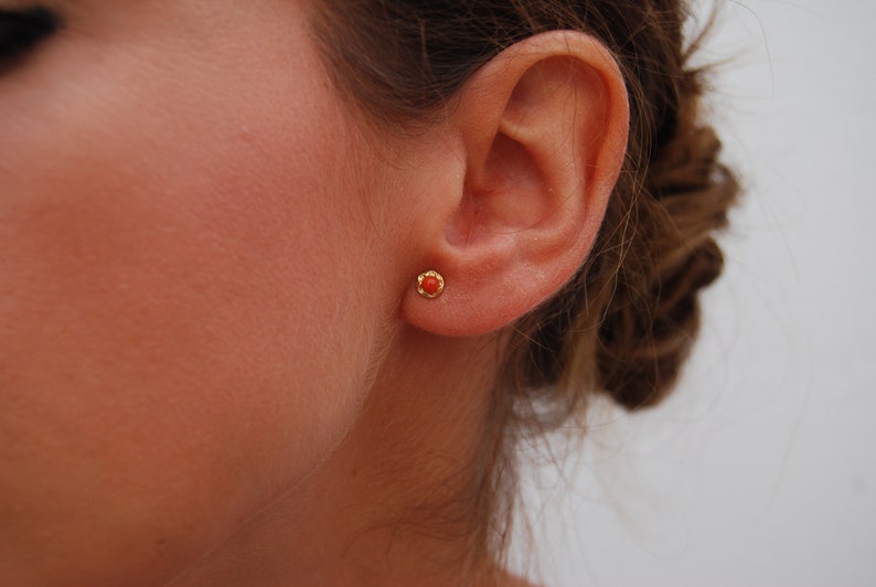 May include: A close-up of a person's ear with a small, red coral stud earring. The earring is set in gold.