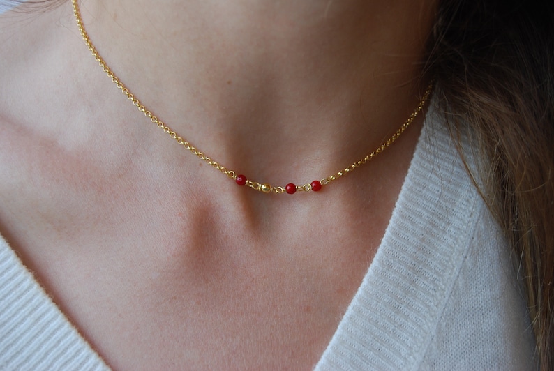 May include: A delicate gold-colored necklace featuring small, round, red beads. The necklace has a thin chain and is worn around the neck. The beads are evenly spaced along the chain, creating a minimalist jewelry design.