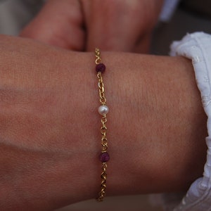 Op de afbeelding: Een gouden kettingarmband met een enkele witte parel en twee kleine robijnrode edelstenen.