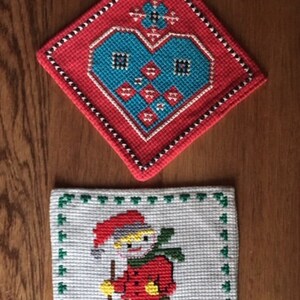 May include: Two handmade, cross-stitched pot holders. The top one is red with a blue heart design. The bottom one is white with a skier in a red jacket and green pants. Both have a decorative border.