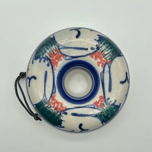 May include: A ceramic donut-shaped object with a central hole and a black cord. The object is white with blue, green, and red painted designs. The inner circle is blue.