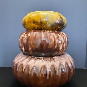 May include: Stacked ceramic bowls in shades of brown and yellow. The top bowl is yellow, the middle bowl is brown with vertical streaks, and the bottom bowl is brown with a lighter, dripped design. The bowls have a rounded shape.