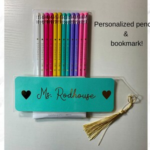 Puede incluir: Un juego de l&aacute;pices personalizados y un marcap&aacute;ginas. Los l&aacute;pices est&aacute;n en una funda de pl&aacute;stico transparente, con una variedad de colores. El marcap&aacute;ginas turquesa tiene el nombre "Ms. Rodhouse" en letras doradas, con un coraz&oacute;n a cada lado. Una borla dorada cuelga.