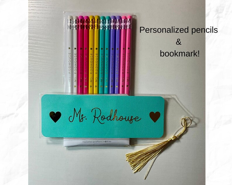 Puede incluir: Un juego de l&aacute;pices personalizados y un marcap&aacute;ginas. Los l&aacute;pices est&aacute;n en una funda de pl&aacute;stico transparente, con una variedad de colores. El marcap&aacute;ginas turquesa tiene el nombre "Ms. Rodhouse" en letras doradas, con un coraz&oacute;n a cada lado. Una borla dorada cuelga.