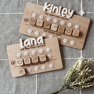 May include: Two wooden morning routine boards with the names "Kinley" and "Lana" in white letters. Each board has a series of icons and the text "my morning routine" and "not needed today". The boards are on a gray patterned surface.